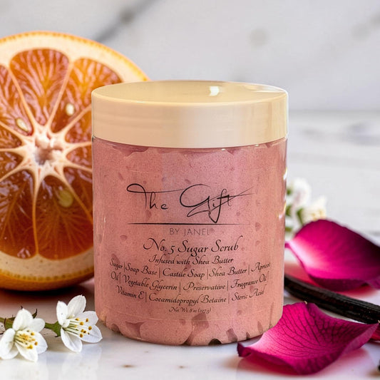 A jar of No. 5 Sugar Scrub by The Gift by Janel styled with sliced oranges, vanilla beans, white flowers, and pink petals on a white marble surface.