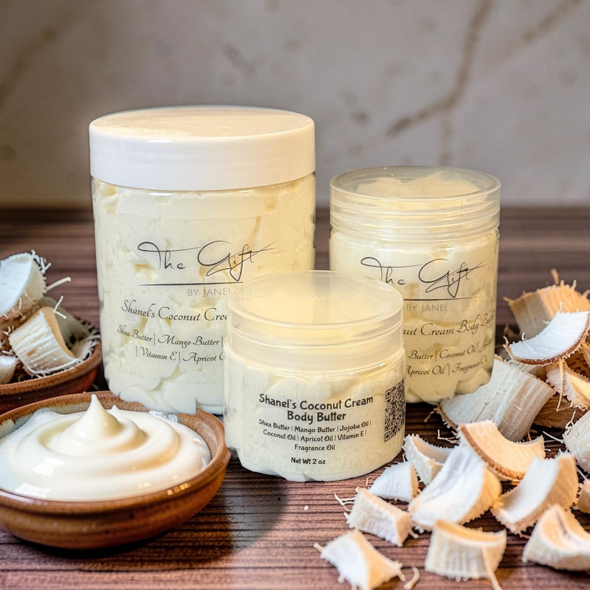 Three jars of Shanel's Coconut Cream Body Butter from The Gift by Janel are arranged on a wooden surface with coconut pieces and a small bowl of cream. The products are shown in a styled setting with a marble background.