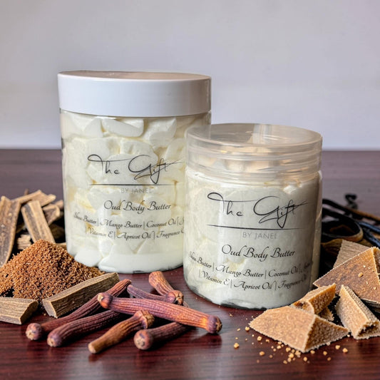 Two jars of The Gift by Janel Oud Body Butter arranged on a dark wooden table with dried wood pieces. The background is a soft, neutral gray gradient.