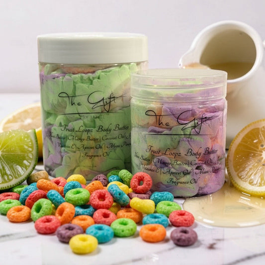 Two jars of 'The Gift' Fruit Loops body butter are displayed on a white marble surface with colorful cereal pieces scattered around them, garnished with lime and orange slices, and a small pitcher of yellow body butter oil in the background.