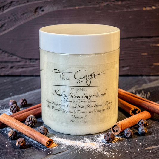 Jar of 'The Gift' by Janel Fluidity Silver Sugar Scrub on a wooden surface with cinnamon sticks and berries.