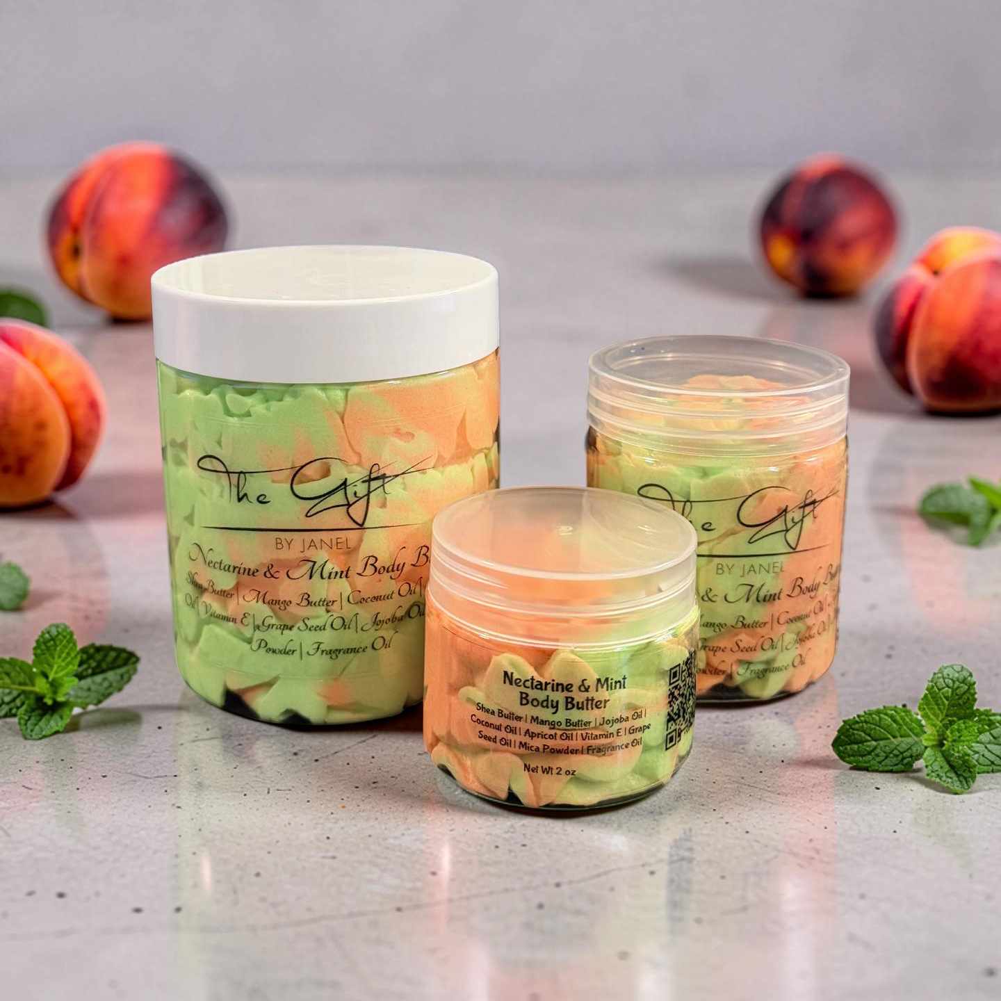 Three jars of 'The Gift' body butter with peaches and mint leaves on a gray surface.