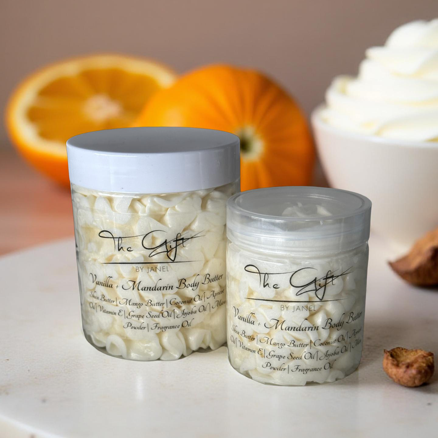 Two jars of The Gift by Janel Vanilla + Mandarin body butter on a marble surface with oranges and vanilla cream in the background.