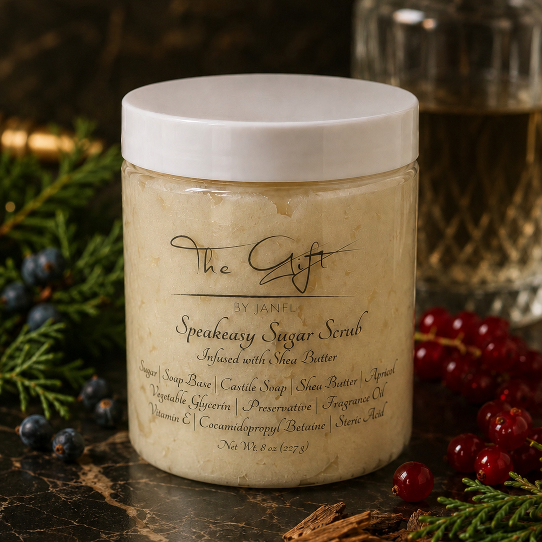 A jar of The Gift by Janel Speakeasy Sugar Scrub sits on a dark marble surface decorated with blueberries, cinnamon, and evergreen branches. The jar has a white lid and a cream-colored scrub inside, with a blurred glass bottle in the background.