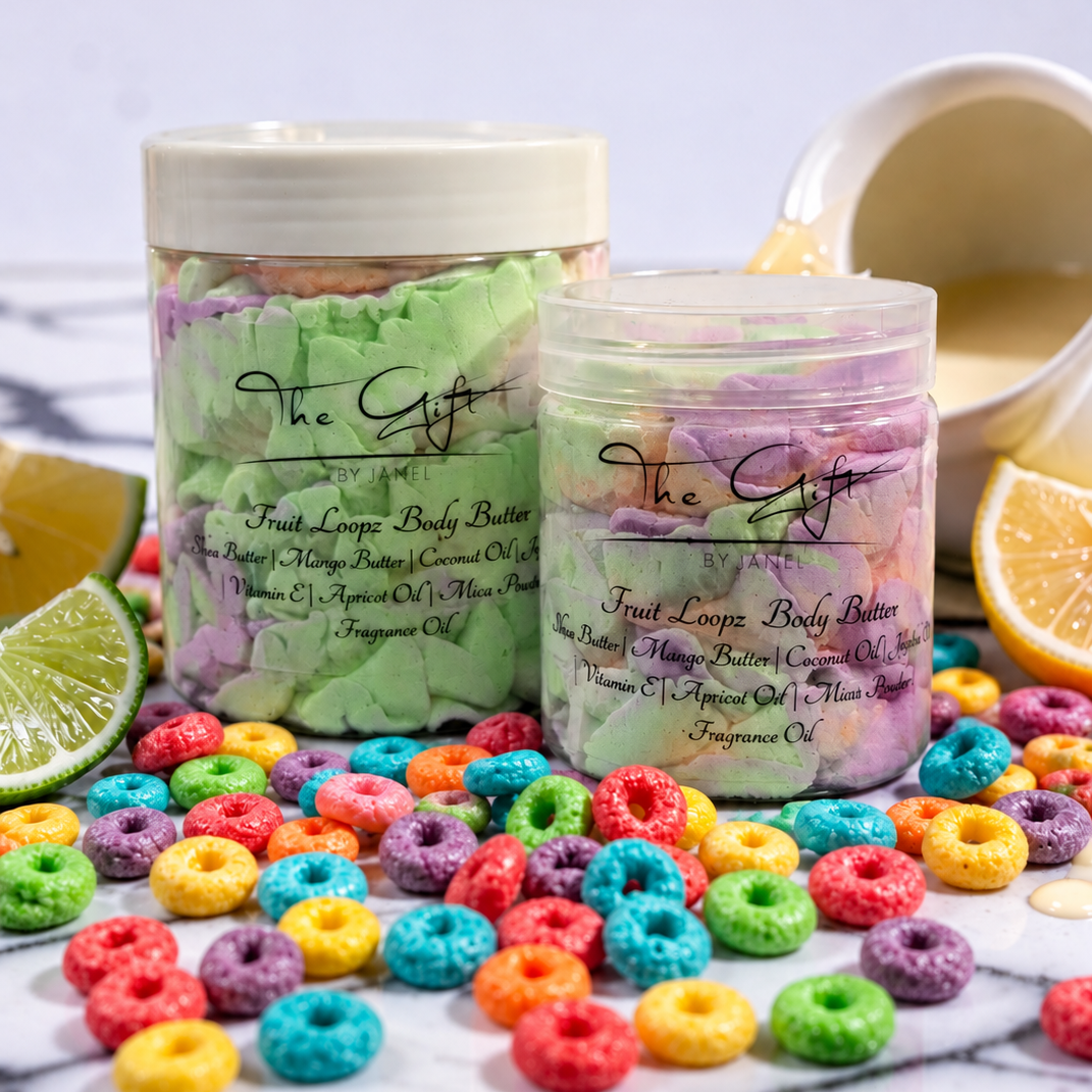 Two jars of 'The Gift' Fruit Loops body butter are displayed on a white marble surface with colorful cereal pieces scattered around them, garnished with lime and orange slices, and a small pitcher of yellow body butter oil in the background.