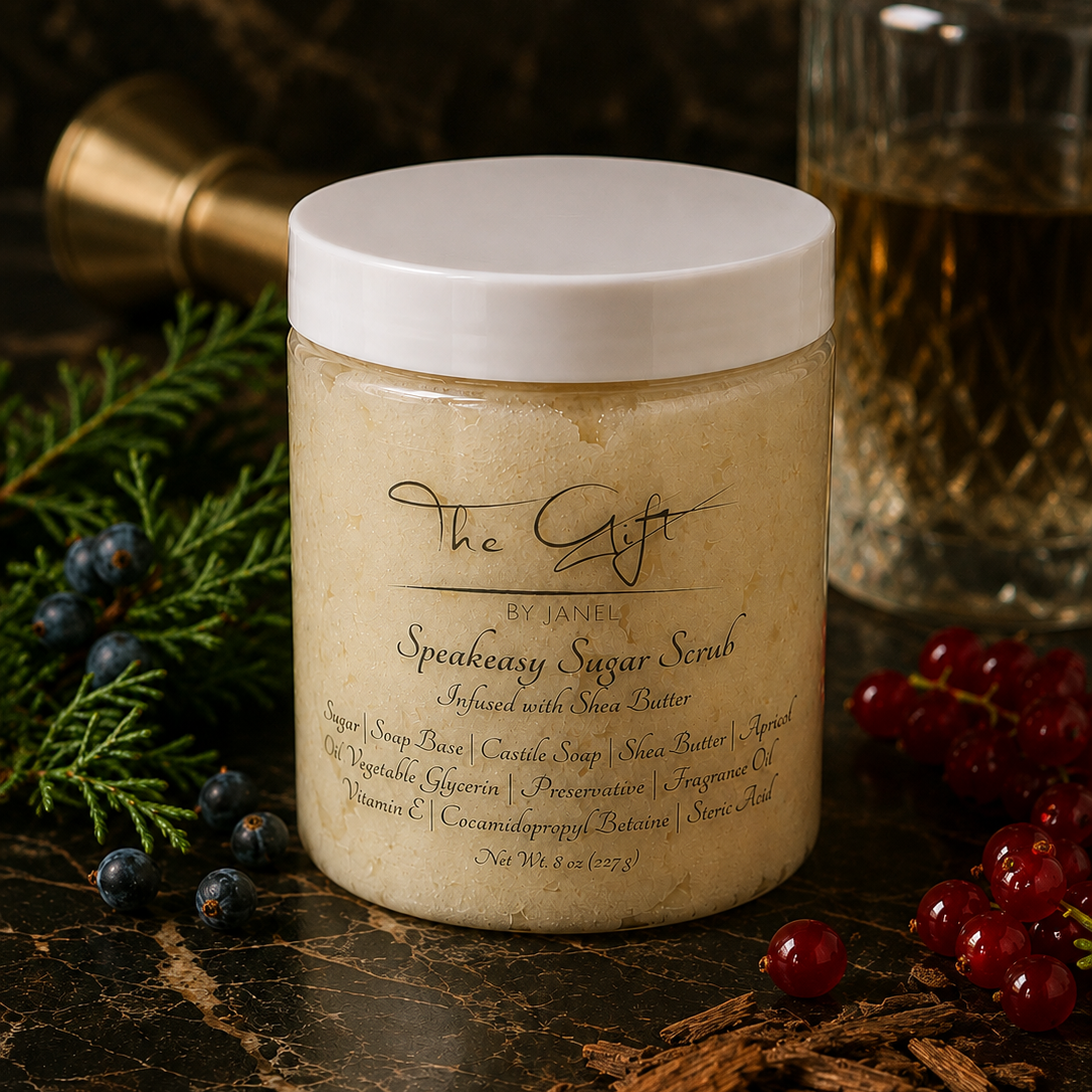A jar of The Gift by Janel Speakeasy Sugar Scrub sits on a dark marble surface decorated with blueberries, cinnamon, and evergreen branches. The jar has a white lid and a cream-colored scrub inside, with a blurred glass bottle in the background.