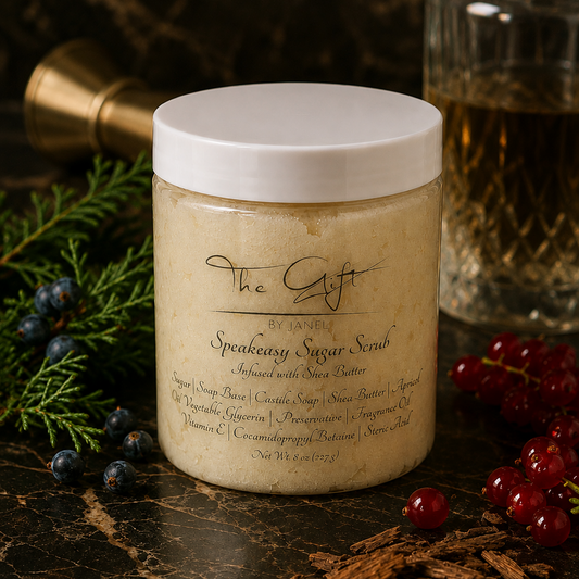 A jar of The Gift by Janel Speakeasy Sugar Scrub sits on a dark marble surface decorated with blueberries, cinnamon, and evergreen branches. The jar has a white lid and a cream-colored scrub inside, with a blurred glass bottle in the background.