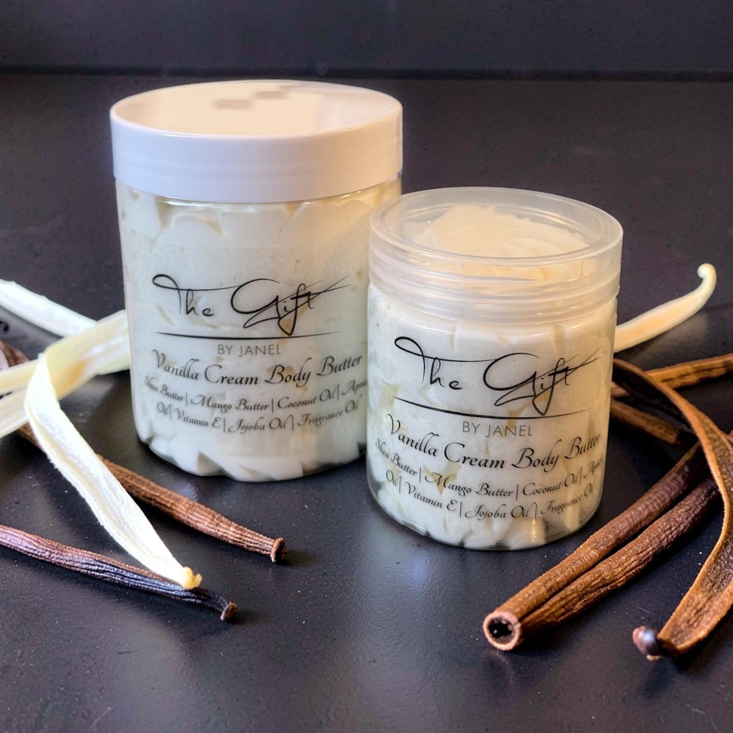 Two containers of Vanilla Cream Body Butter with vanilla beans on a dark surface