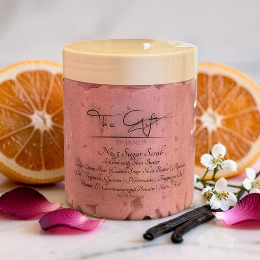 A jar of No. 5 Sugar Scrub by The Gift by Janel styled with sliced oranges, vanilla beans, white flowers, and pink petals on a white marble surface.