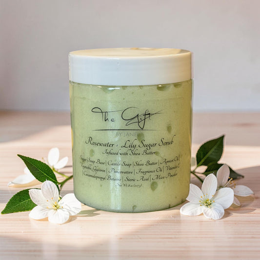 A jar of 'The Gift' by Janel 'Rosewater + Lily Sugar Scrub' sits on a light wooden surface, surrounded by white blossoms and green leaves. The scrub inside is a pale, yellowish-green, and the jar has a white lid.