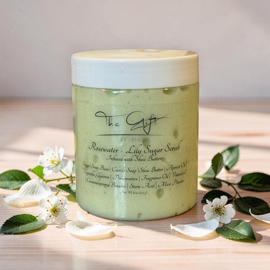 A jar of 'The Gift' by Janel Rosewater + Lily Sugar Scrub sits on a light wooden surface surrounded by white blossoms and green leaves. The scrub inside is a pale, yellow-green color, and the jar has a white lid.
