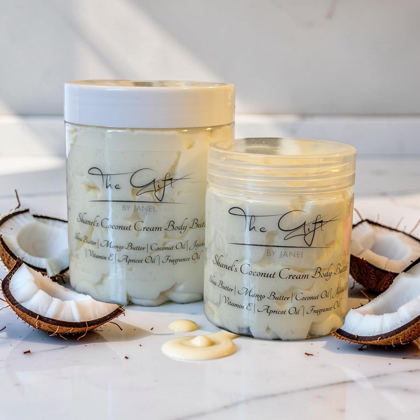 Two jars of Shanel's Coconut Cream Body Butter by The Gift by Janel are displayed on a white marble surface with sliced coconuts and dollops of the cream-colored product around them.
