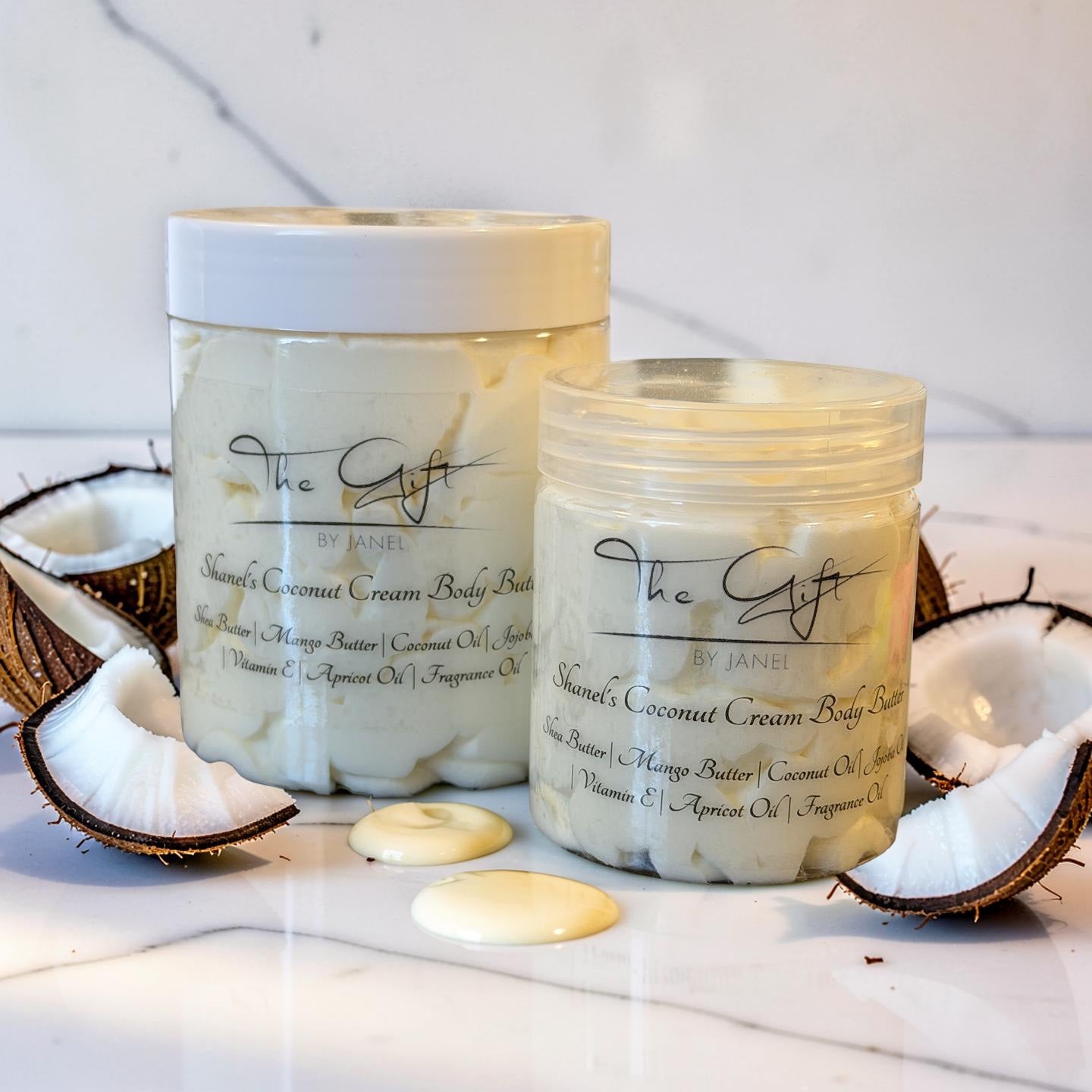 Two jars of Shanel's Coconut Cream Body Butter by The Gift by Janel are displayed on a white marble surface with sliced coconuts and dollops of the cream-colored product around them.