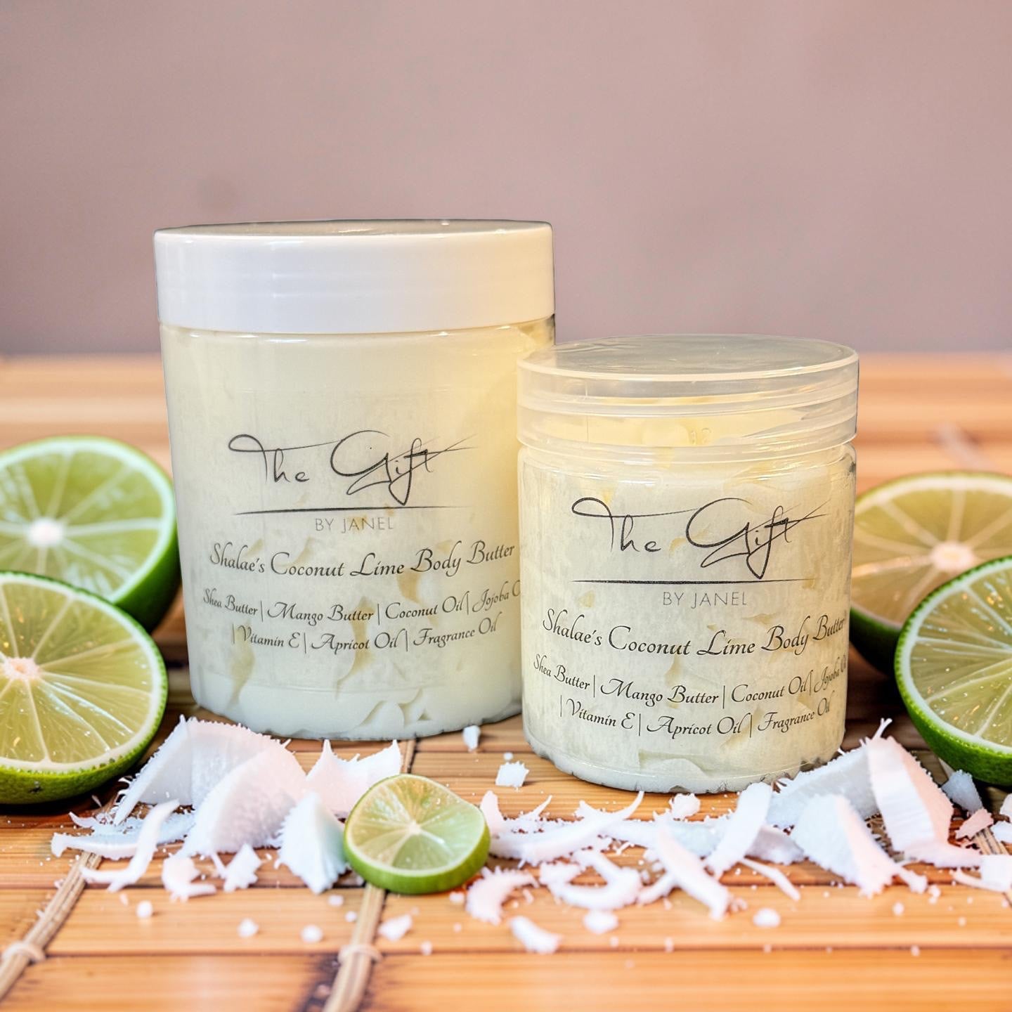 Two jars of 'The Gift by Janel natural coconut lime body butter set on a wooden board, surrounded by lime slices, coconut flakes, and shredded coconut against a neutral background.