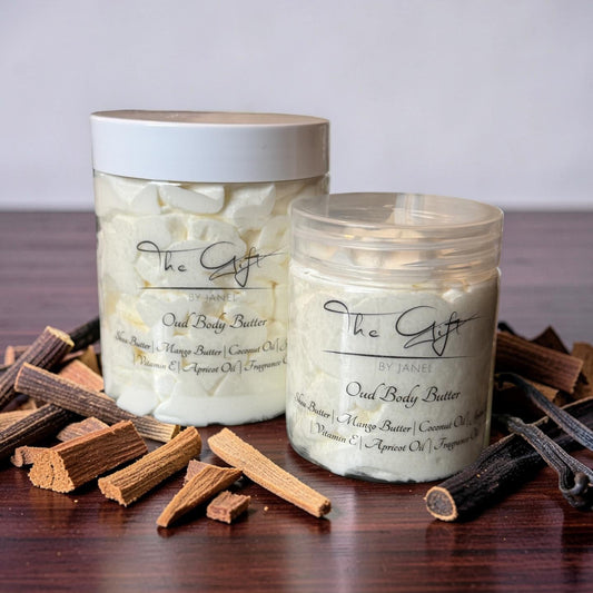 Two jars of The Gift by Janel Oud Body Butter arranged on a dark wooden table with dried wood pieces. The background is a soft, neutral gray gradient.