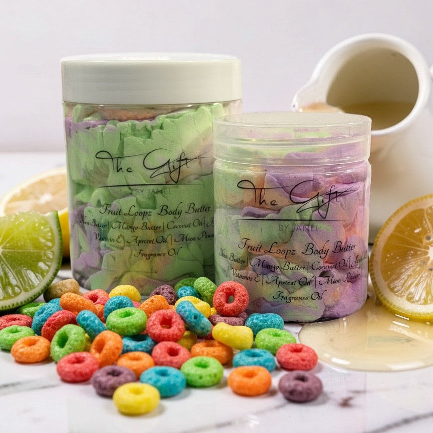 Two jars of 'The Gift' Fruit Loops body butter are displayed on a white marble surface with colorful cereal pieces scattered around them, garnished with lime and orange slices, and a small pitcher of yellow body butter oil in the background.