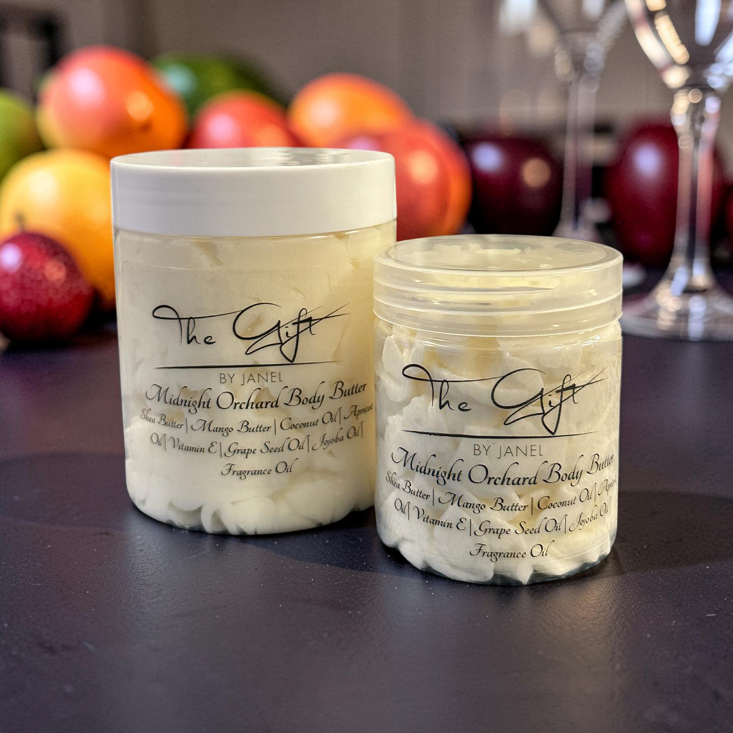Two containers of 'The Gift' by Janel Midnight Orchard Body Butter on a dark surface with apples in the background.
