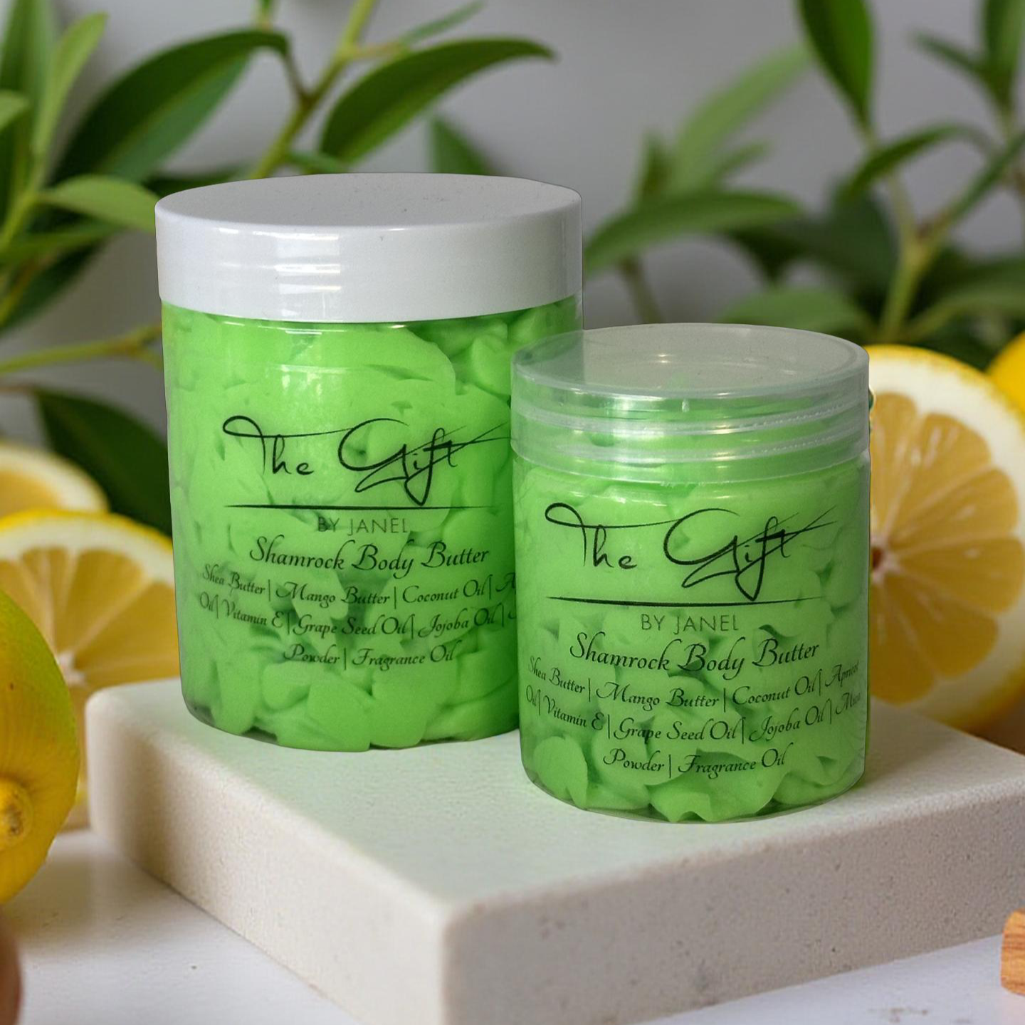 Two green jars of The Gift by Janel Shamrock Body Butter on a white surface with lemons and green leaves in the background.