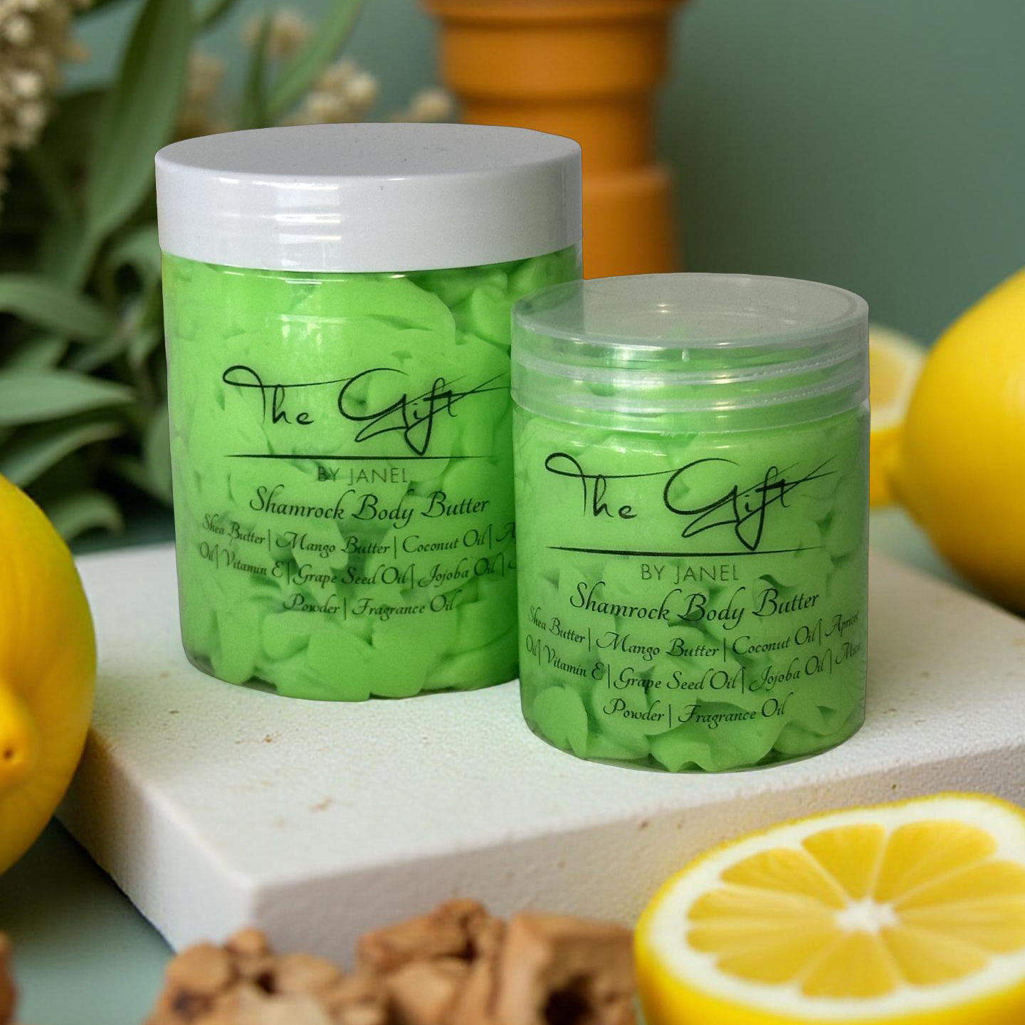 Two jars of The Gift by Janel Shamrock Body Butter on a surface with lemons and woods.