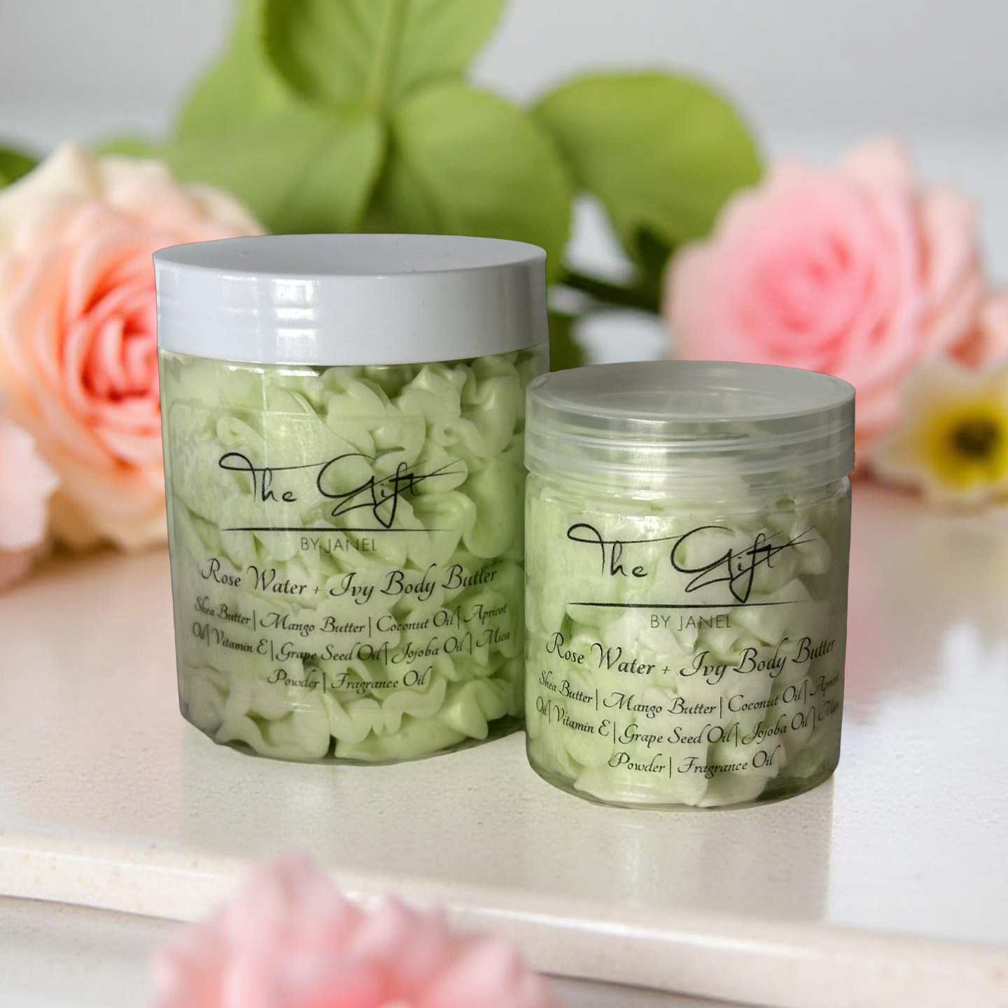 Two jars of body butter labeled The Gift by Janel on a surface with flowers in the background.